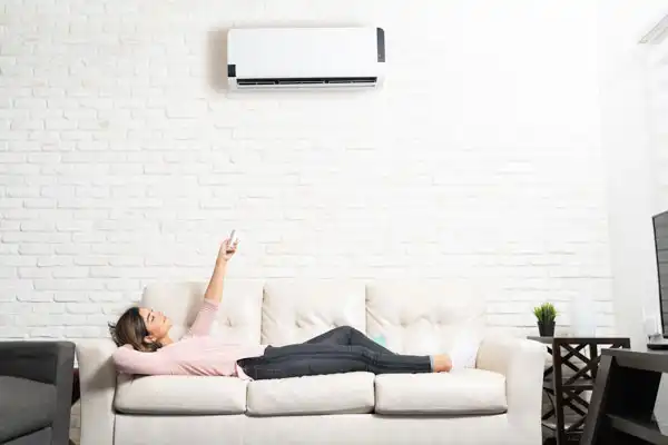 Person lying on a sofa in a white room, using a remote control to operate an air conditioner mounted on the wall above.