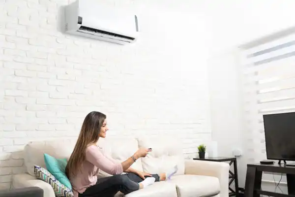 Woman sitting on a couch in a bright room, using a remote control. Air conditioner on the wall and a television nearby.