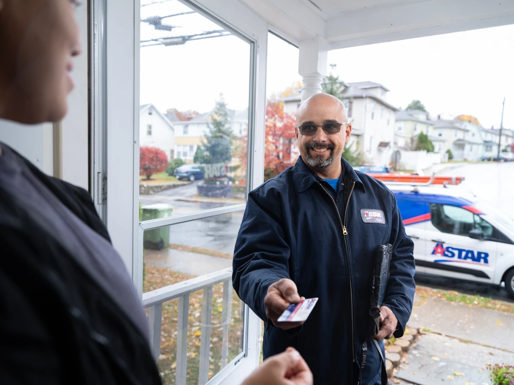 A delivery worker hands a card to a person at a front door; a company van marked “ASTAR” is parked outside on a rainy day.