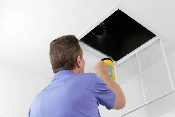 Person in a blue shirt shining a flashlight into an open ceiling vent.