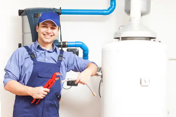 Plumber in blue overalls holding a wrench and pipe, standing next to a water heater.