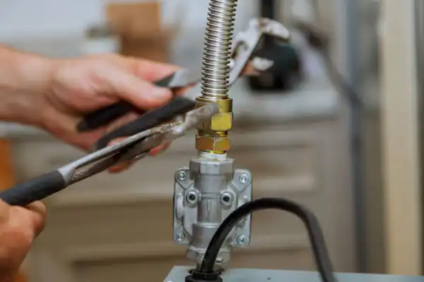 Hands using pliers to tighten a metal gas fitting connected to a flexible pipe, with tools and machinery visible in the background.