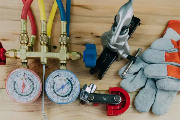 Tools on a wooden surface include a manifold gauge set, pipe cutter, gloves, and a flaring tool, used for HVAC or plumbing tasks.