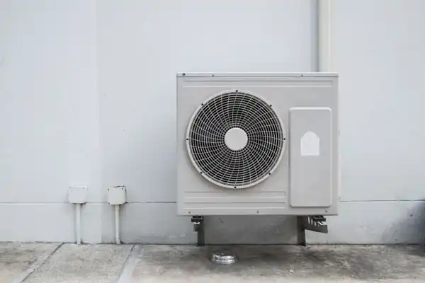 Outdoor air conditioning unit mounted on a wall, with pipes and electrical conduits visible nearby.