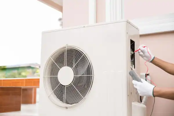A technician wearing gloves uses a multimeter to check an air conditioning unit outdoors.