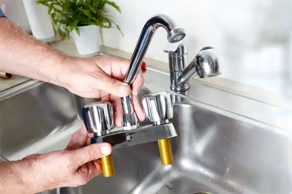 Person holding a detached kitchen faucet with two knobs above a stainless steel sink.