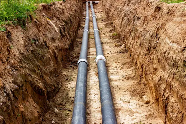 Two black pipes are laid in a trench dug into the earth, surrounded by soil and grass.