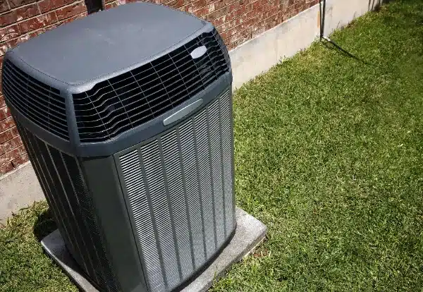 An outdoor air conditioning unit on a concrete base next to a brick wall, surrounded by grass.