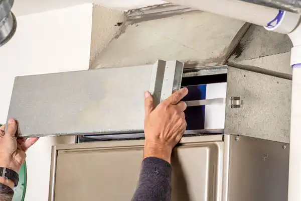 Person removing a panel from a metal furnace in a building, exposing the air filter inside.
