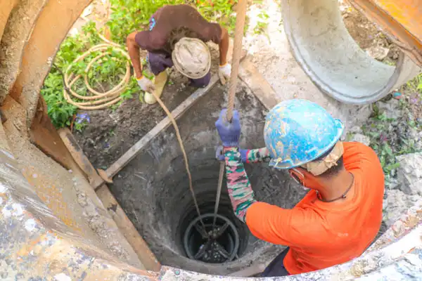 Two construction workers lowering a tool into a deep cylindrical hole with ropes. One wears an orange shirt and blue helmet; the other has a dark shirt and a white helmet.